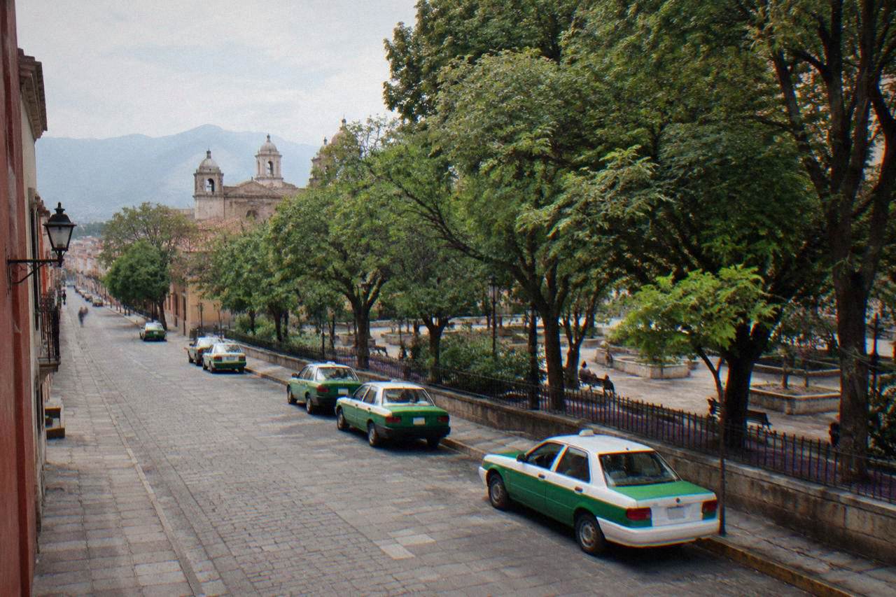 A narrow colonial street in Oaxaca City with green-and-white taxis parked along the curb