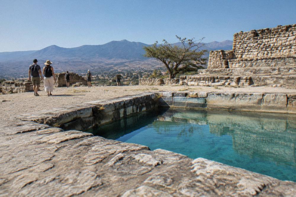 Visitors walking among stone ruins on a dry hilltop in the Oaxaca valleys under a blue sky