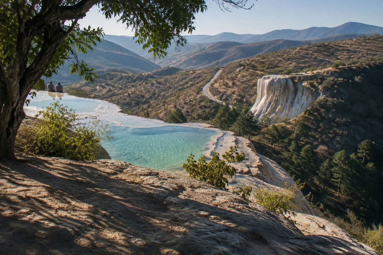 A mountain road winding through dry hills toward the mineral terraces of Hierve el Agua in the distance