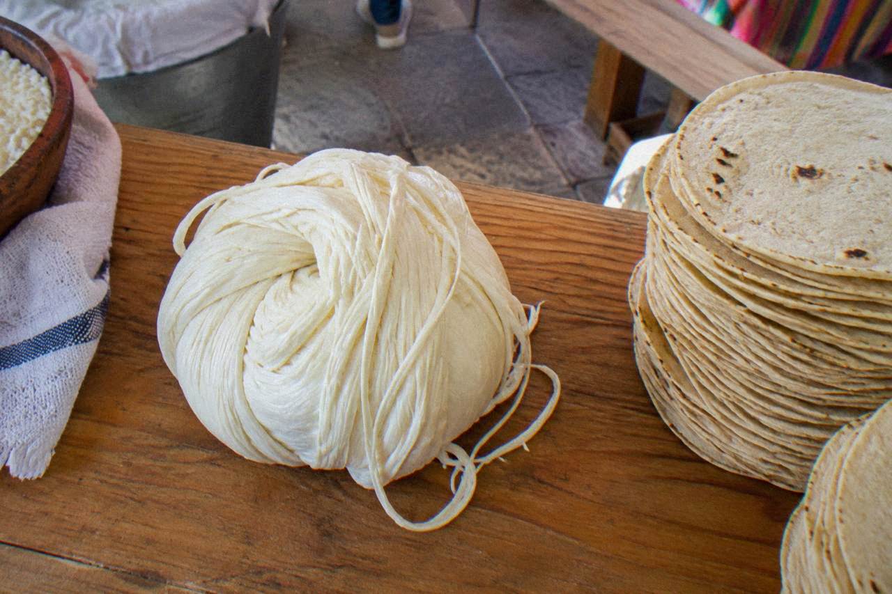 A ball of white string cheese resting on a wooden market counter beside stacked tortillas