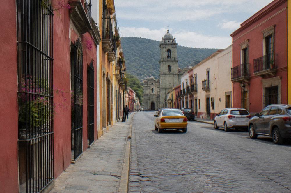 A narrow Oaxaca street with coloured walls and a taxi driving past parked cars