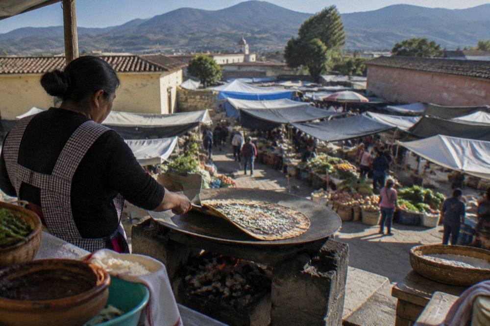 A cook flipping a large tlayuda on a charcoal griddle at an open-air market stall