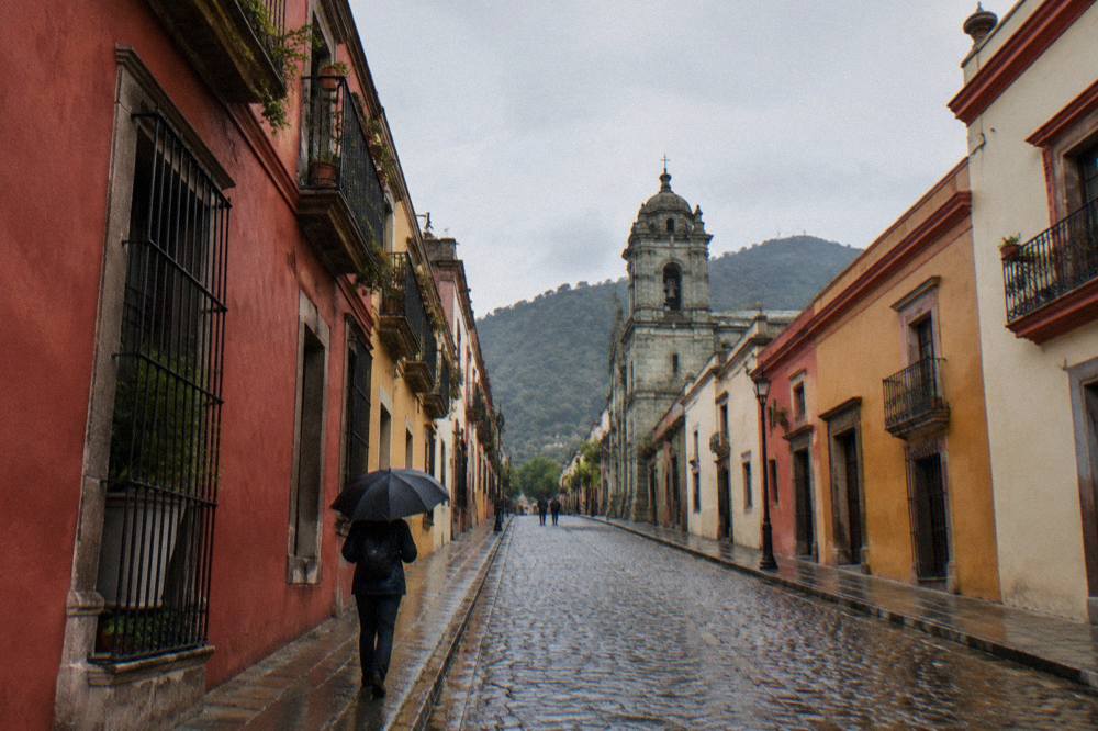A visitor in a rain jacket holding an umbrella on a wet colonial street in Oaxaca City