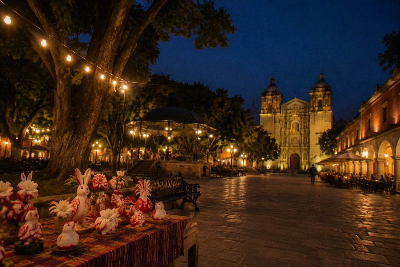 Carved radish figures displayed on tables in a plaza at night, lit by strings of lights