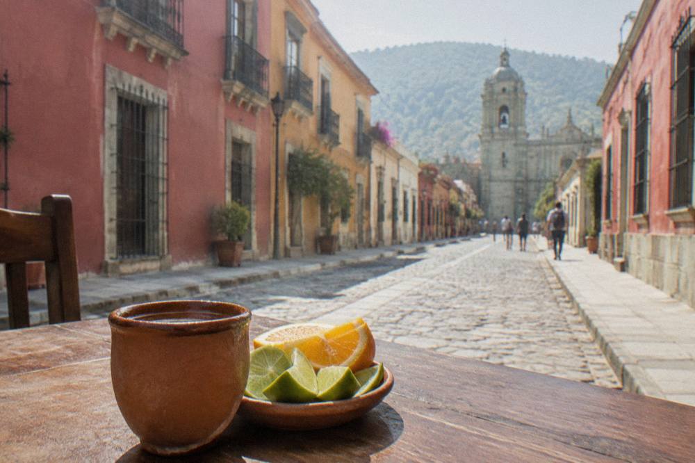 A clay cup of mezcal beside lime wedges and orange slices on a wooden bar