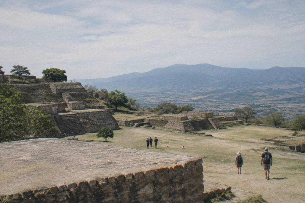 Visitors walking among the stone terraces of the Monte Albán archaeological site on a hillside above a valley
