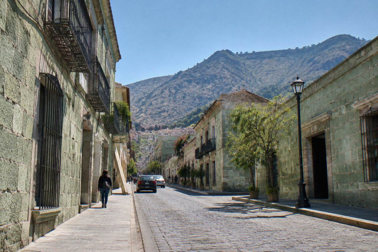 A busy Oaxaca city street with green stone buildings and mountains rising behind the rooftops