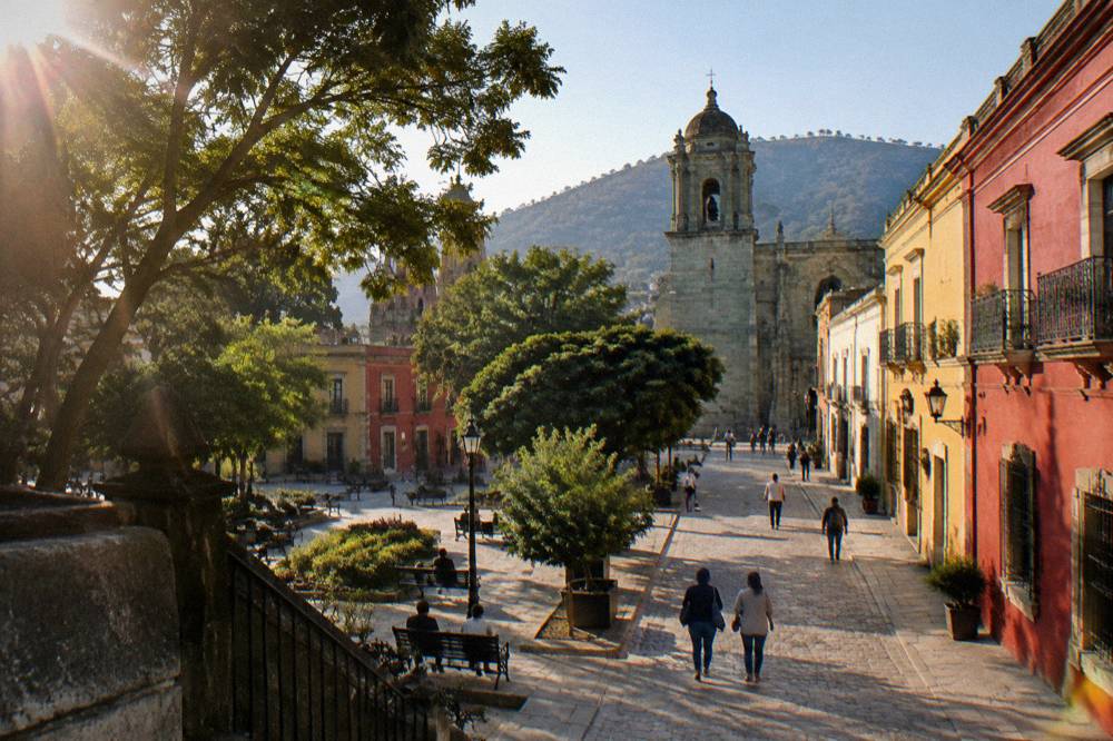 An open plaza in Oaxaca City with trees, benches and people passing through