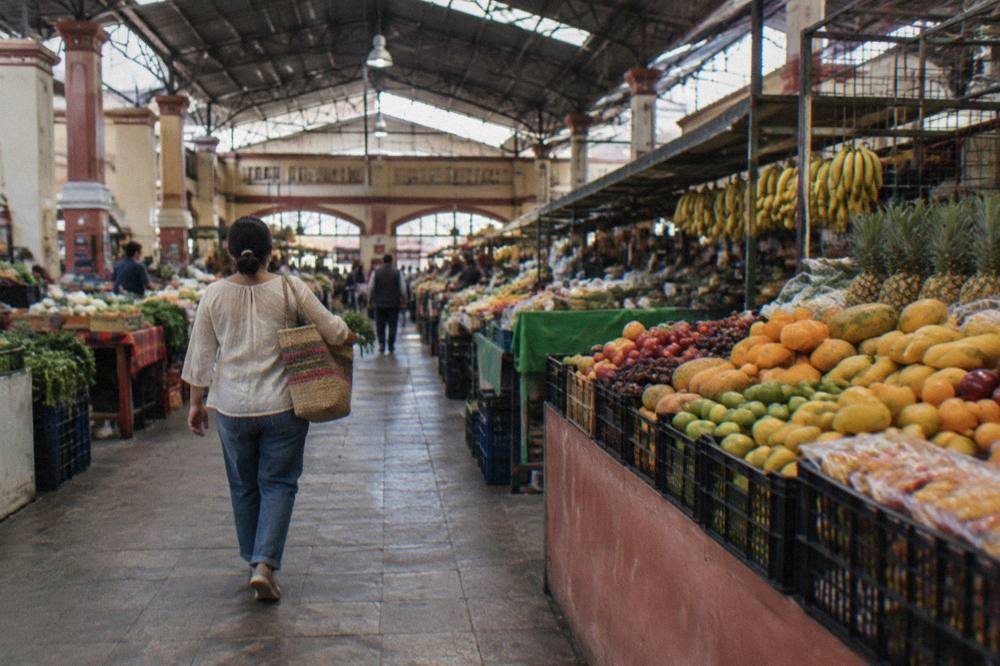 A shopper carrying a woven market bag past fruit stalls in a covered Oaxaca market hall