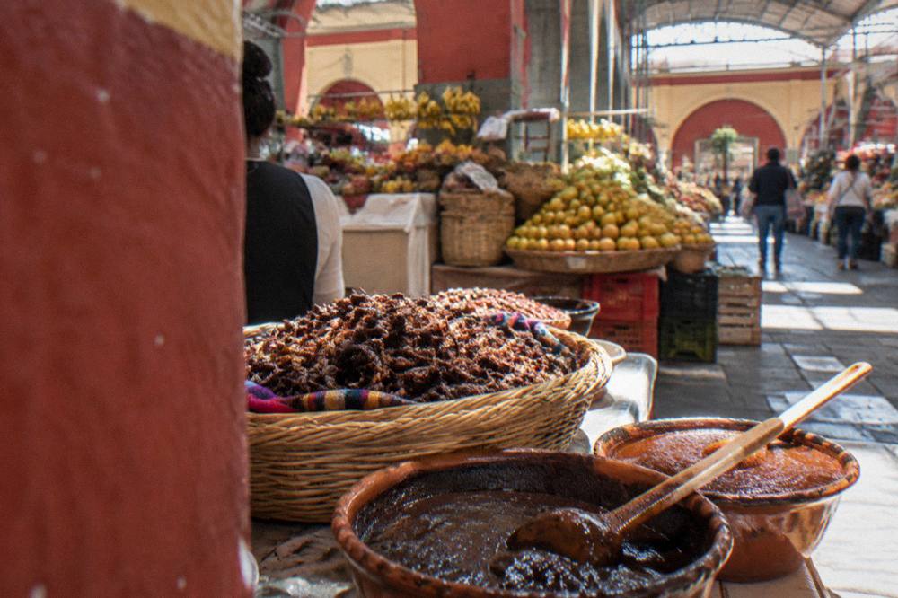 Basket of toasted chapulines beside ladles of mole paste at a market counter
