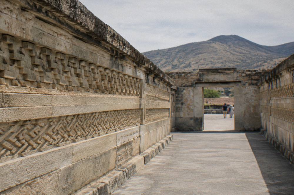 Long corridor of stone walls with repeating carved patterns leading toward an open doorway