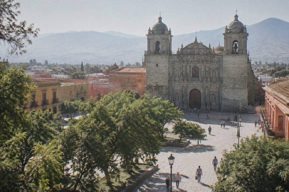 People walking across a paved city square with trees and colonial buildings on all sides