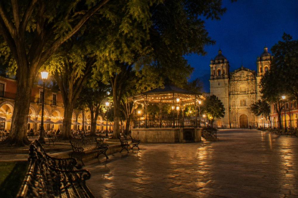 The Zócalo plaza at night, warm lamp light on the stone arcades, laurel trees and empty iron benches under a deep blue evening sky