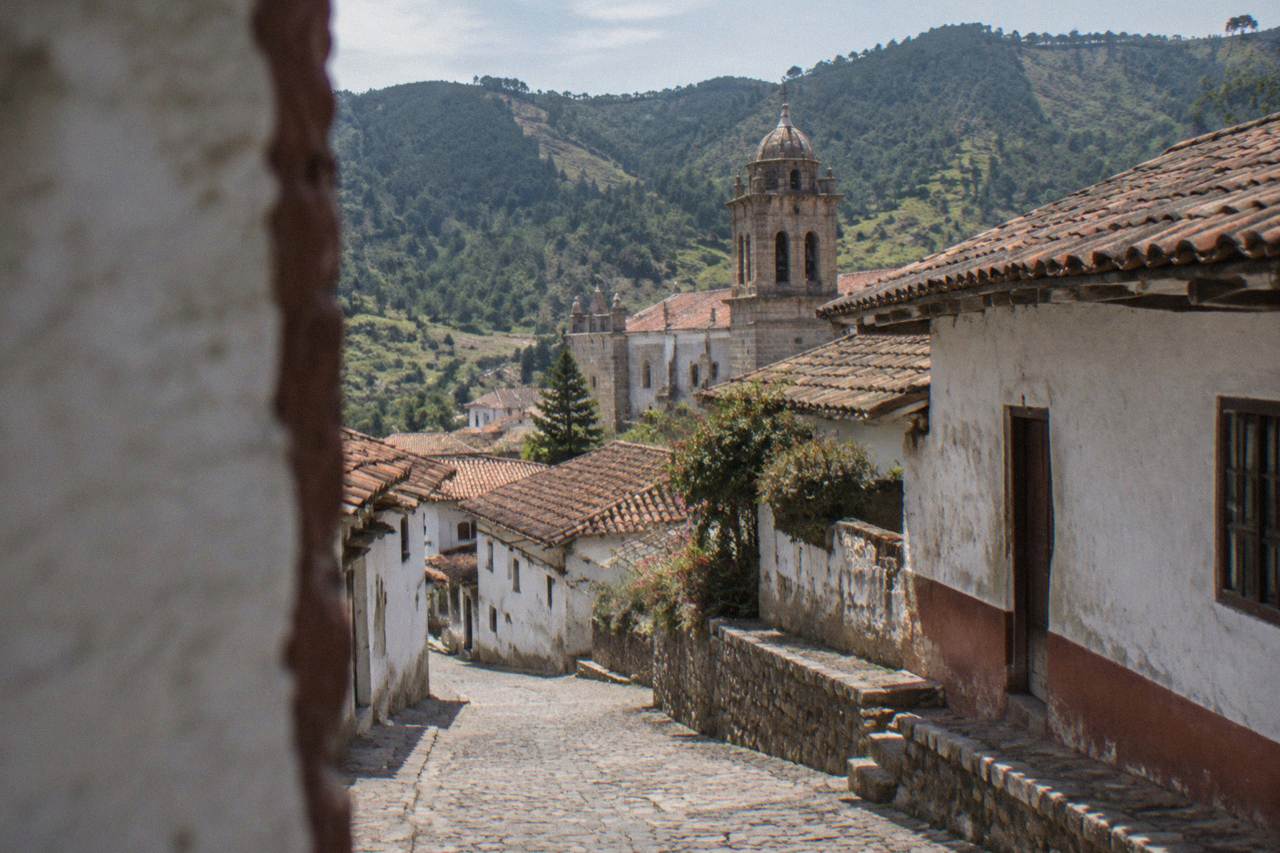 A steep cobbled street of whitewashed houses with red-tile roofs in Capulalpam de Mendez, pine ridges behind