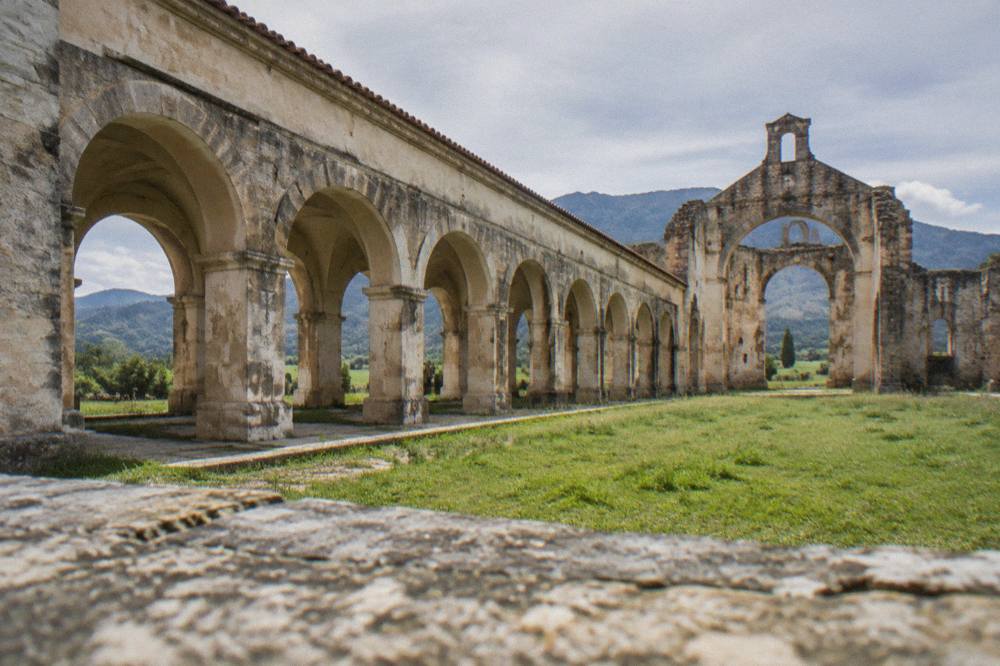 Open stone arches of a monastery nave with grass and sky visible through them