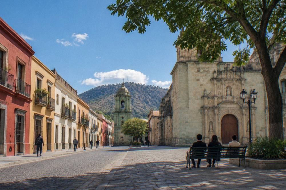 A shaded plaza in Oaxaca City with a church facade and people resting on a bench