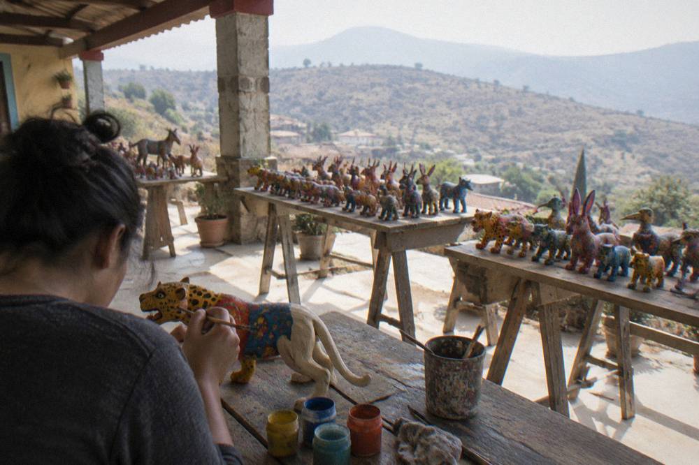 A visitor holding a brush over a half-painted carved jaguar figure at a wooden workbench
