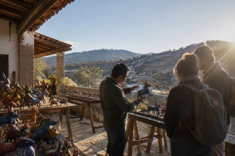 A local artist explaining a painted wooden figure to two visitors inside a small workshop