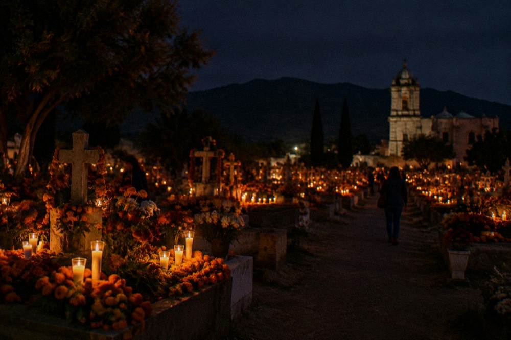A cemetery at night with graves covered in orange marigolds and candles glowing, seen from a distance