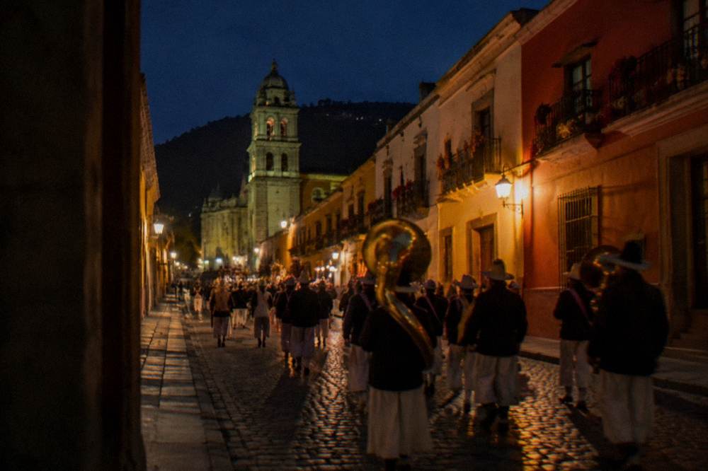 A costumed procession with brass instruments moving down a decorated Oaxaca street at night