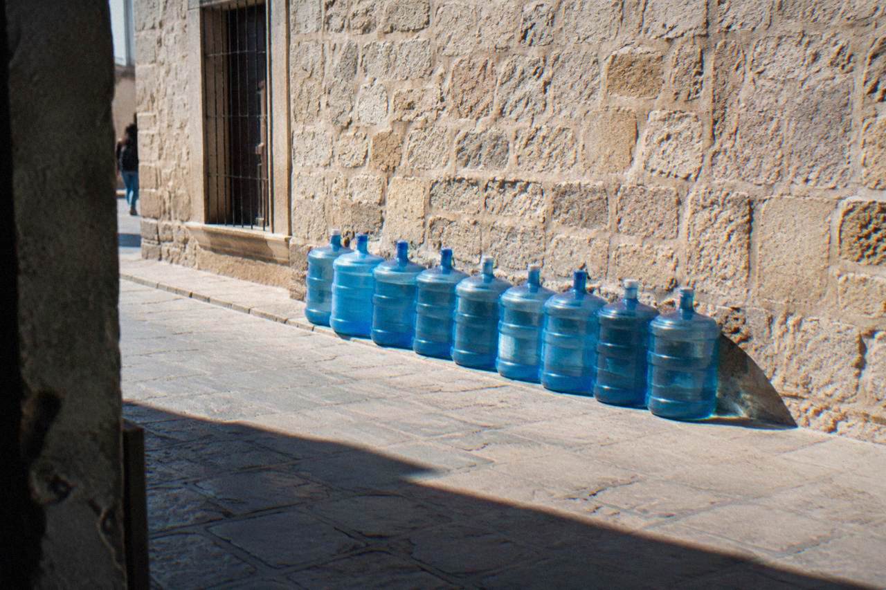 Large blue water jugs lined up against a sunlit colonial stone wall on a street in Oaxaca City