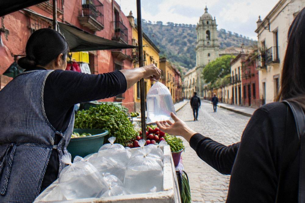 A vendor handing a sealed plastic bag of purified water across a market stall counter in Oaxaca