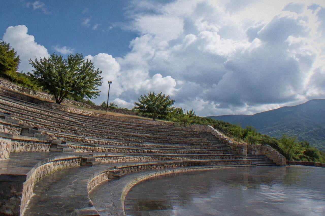 A hillside open-air amphitheater with tiered stone seating and dry grass under a bright summer sky, empty of people