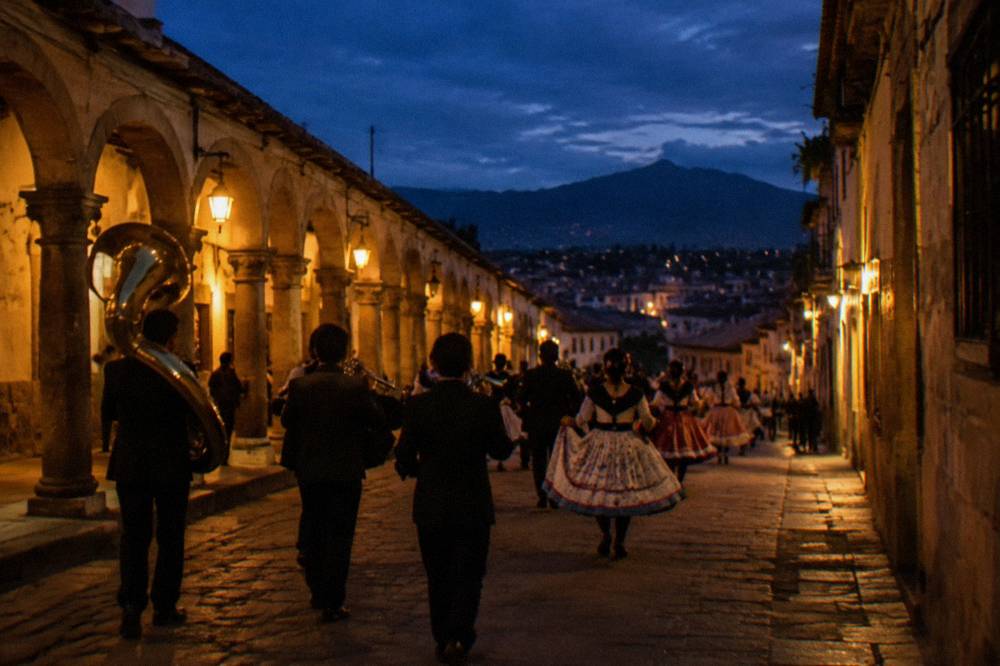 A procession with brass musicians and dancers moving through a colonnaded city street at dusk