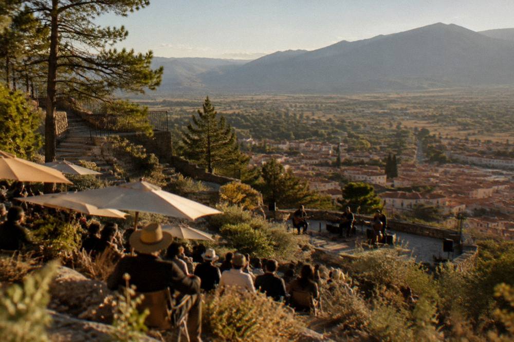 Rows of spectators under parasols on a sunlit hillside facing an open-air stage