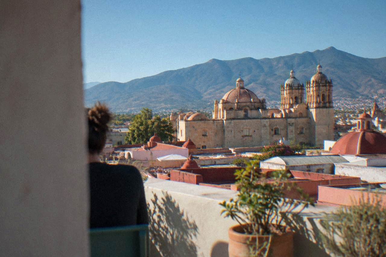 Sunlit rooftops of Oaxaca City church domes under a clear blue sky with green hills behind