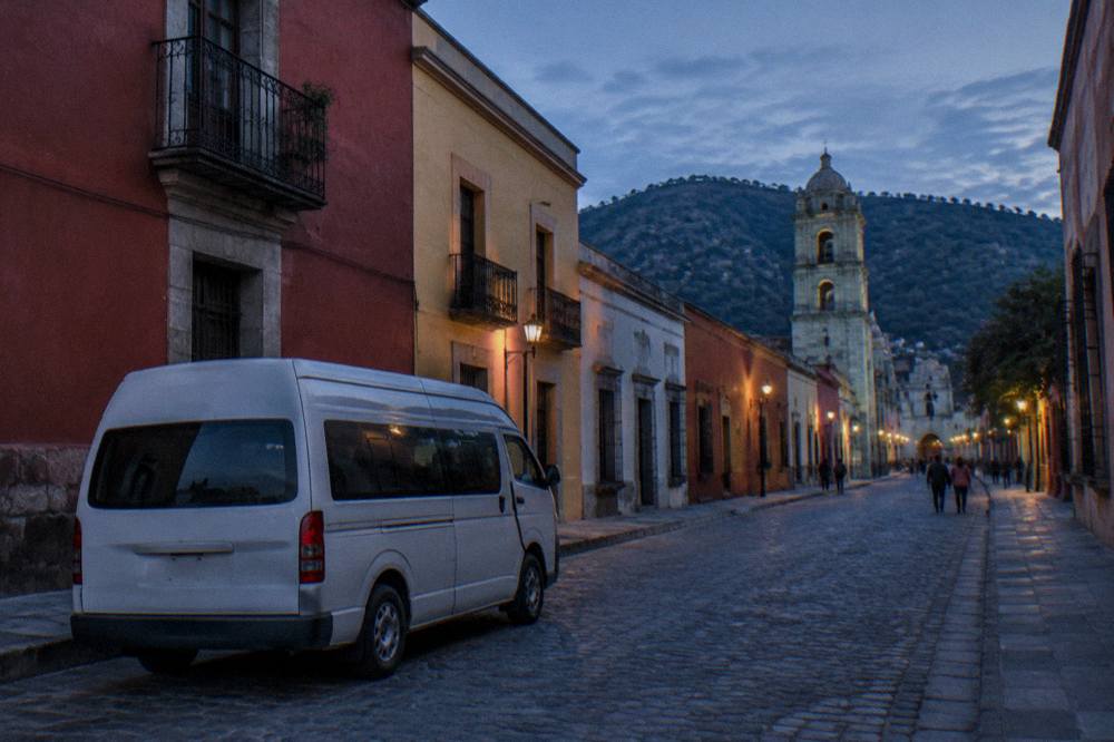 A shared van colectivo parked on a street in Oaxaca with hills behind