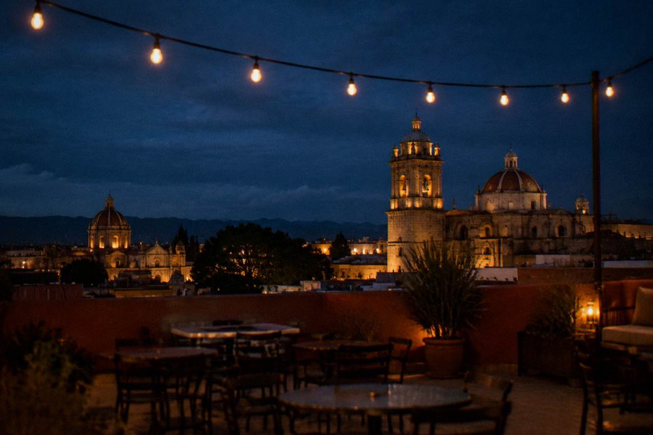 A rooftop terrace at dusk with string lights and empty tables overlooking domes and bell towers of the historic center
