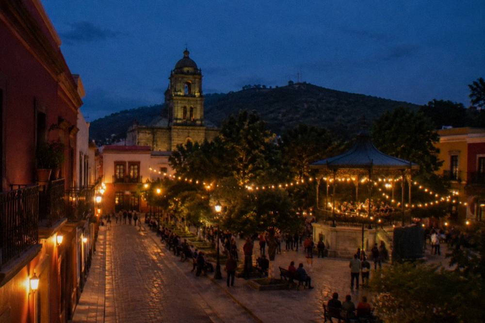 An evening plaza with string lights and people gathered around a bandstand