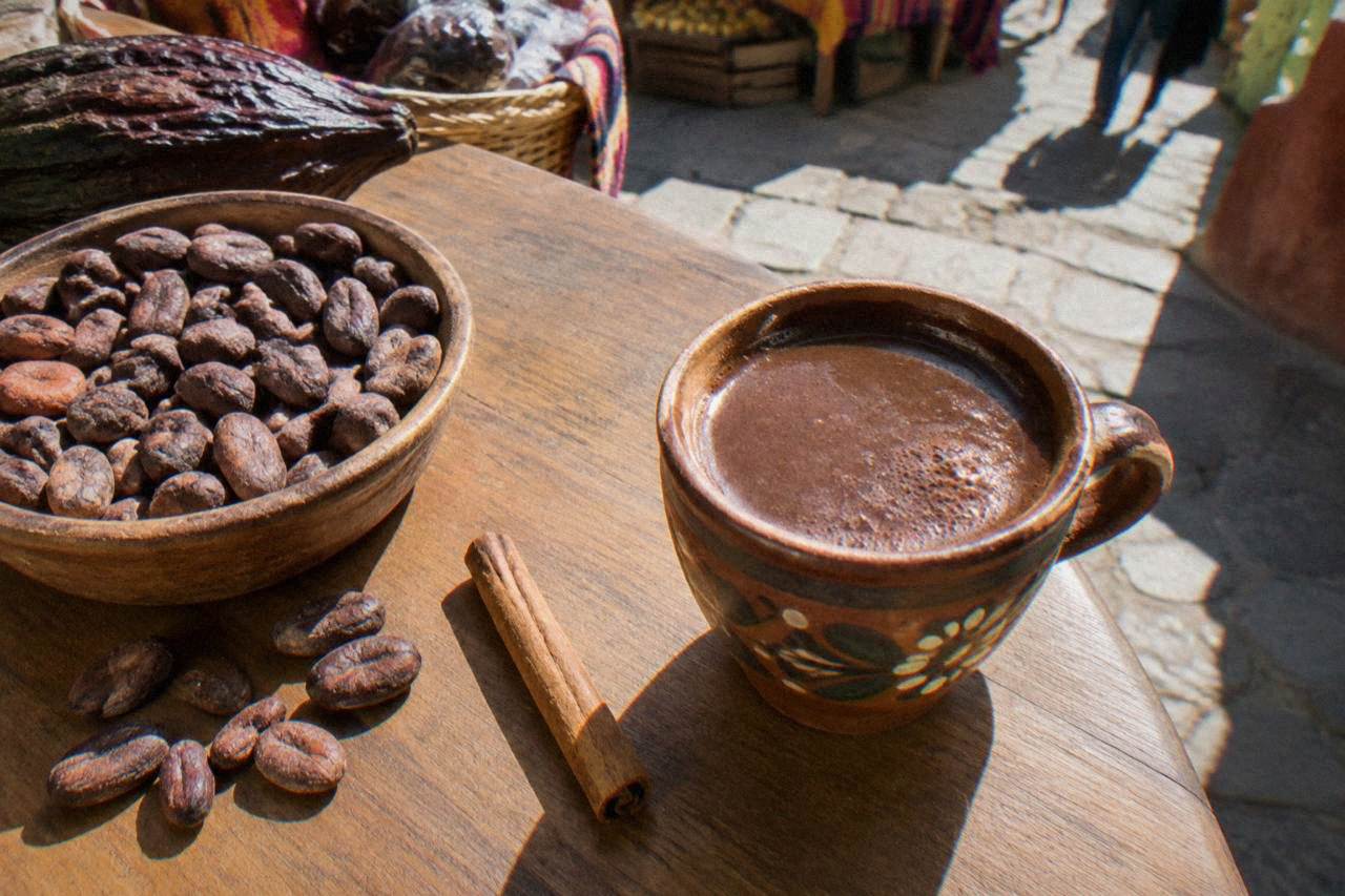 A cup of dark hot chocolate beside cacao beans and a cinnamon stick on a wooden market counter