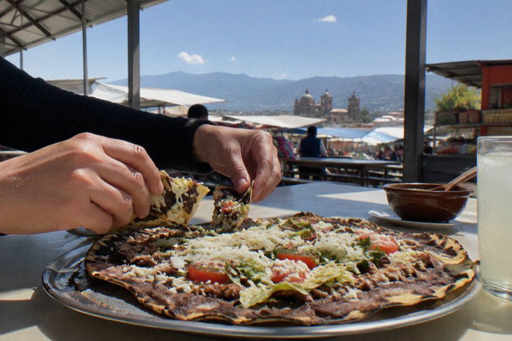 Hands tearing pieces from a large folded tlayuda on a metal tray at an Oaxaca market comedor