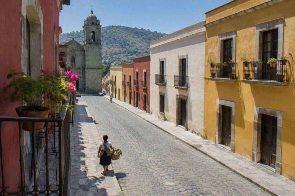 A shopper carrying a basket of fresh vegetables through a sunny Oaxaca street