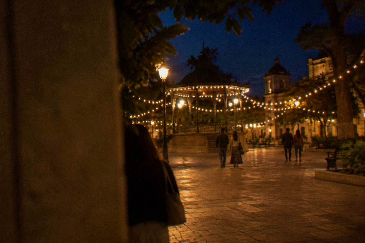A lit plaza at night with strings of warm lights and strollers seen from behind in Oaxaca City