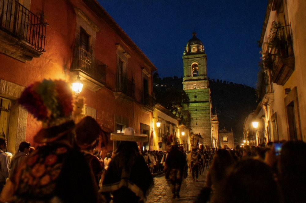 A costumed comparsa procession moving through a crowded lit street in Oaxaca City at night