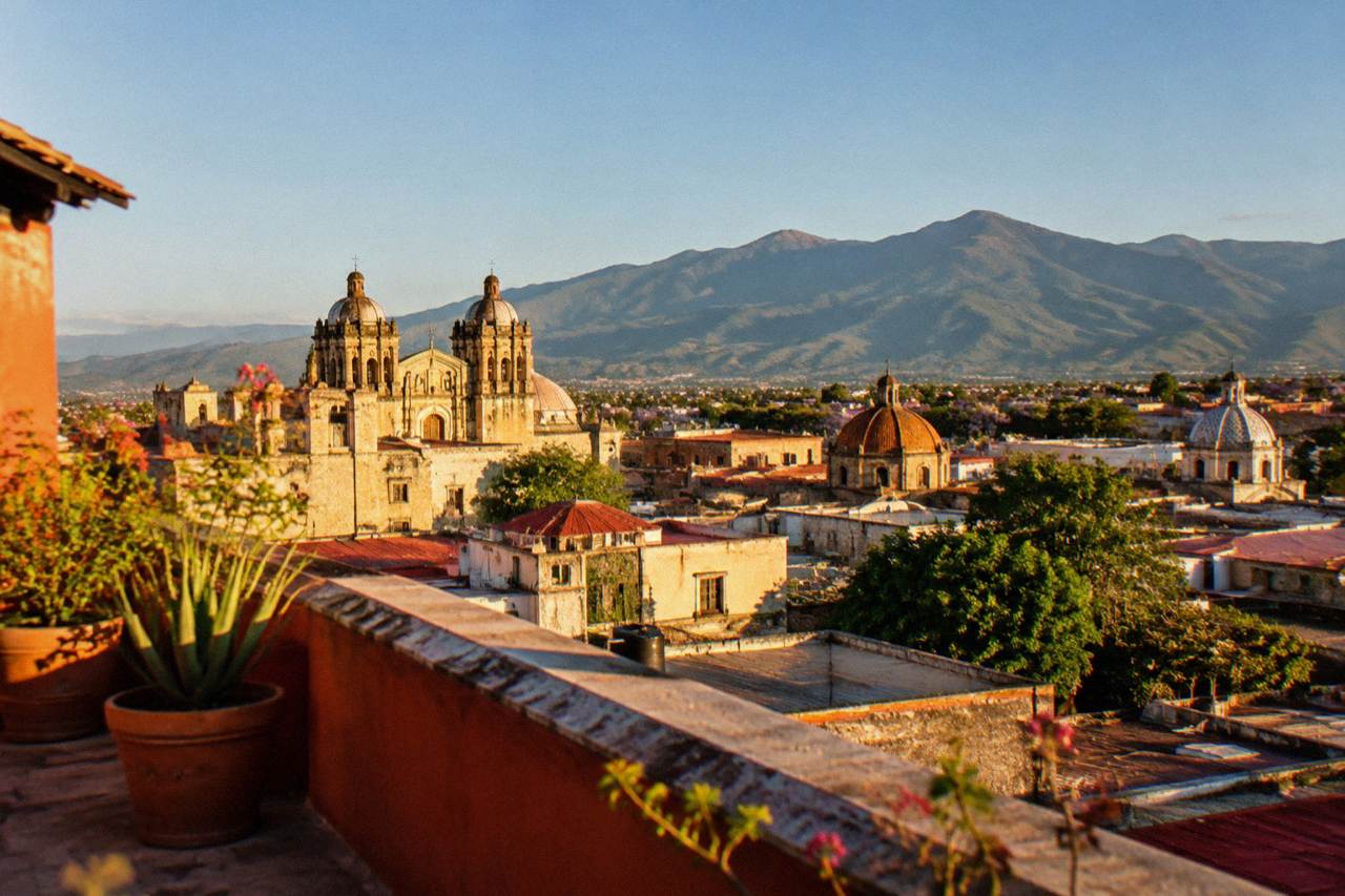 View over the rooftops and church domes of Oaxaca City toward green mountains at midday