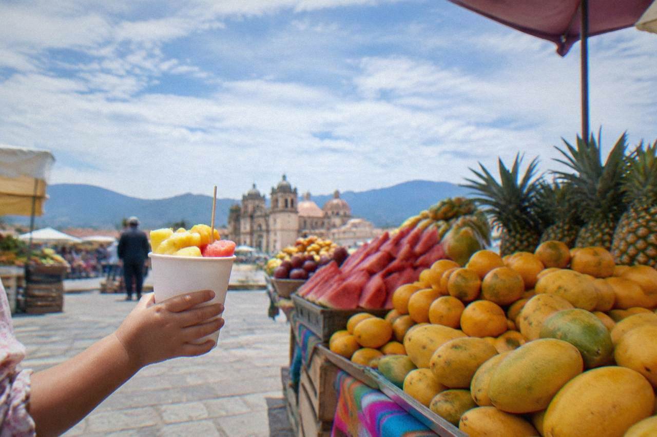 A child's hand holding a paper cup of cut fruit at a market stall in Oaxaca