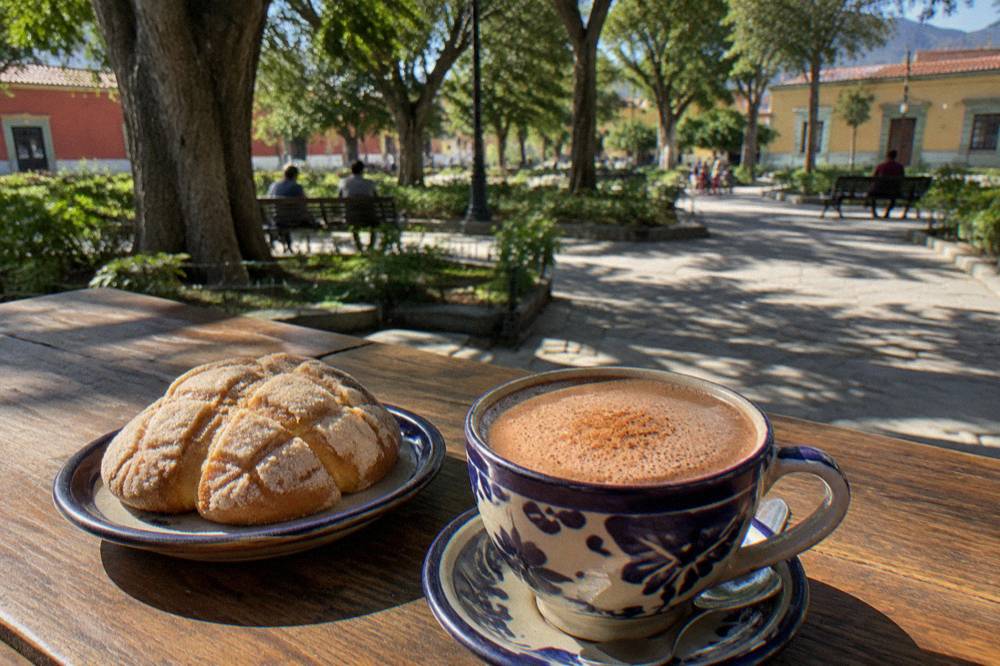 A cup of frothed Oaxacan hot chocolate beside sweet bread on a wooden table