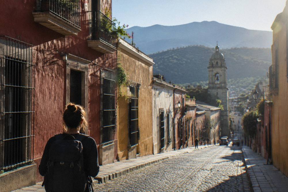 A solo traveller with a backpack looking down a colourful Oaxaca street toward distant mountains