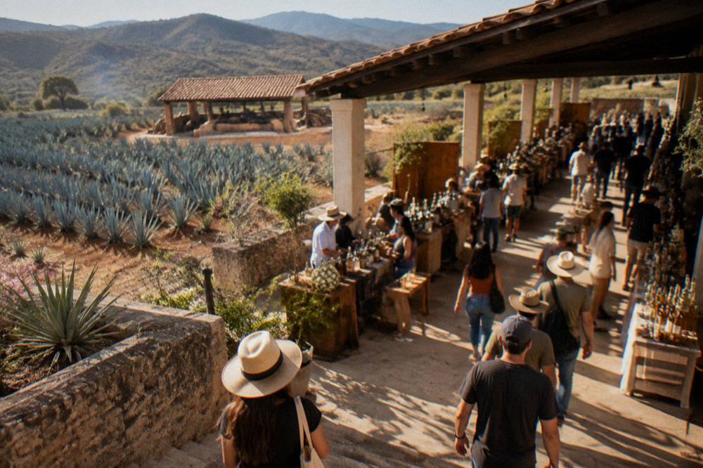 Crowds walking through a mezcal fair hall with rows of producer stands and bottled mezcal